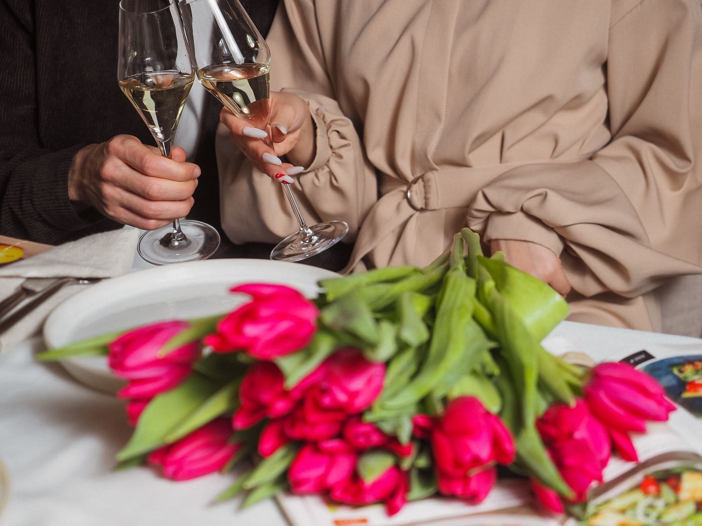 A dinner table set for two, hands raising wine glasses beside a bunch of pink tulips